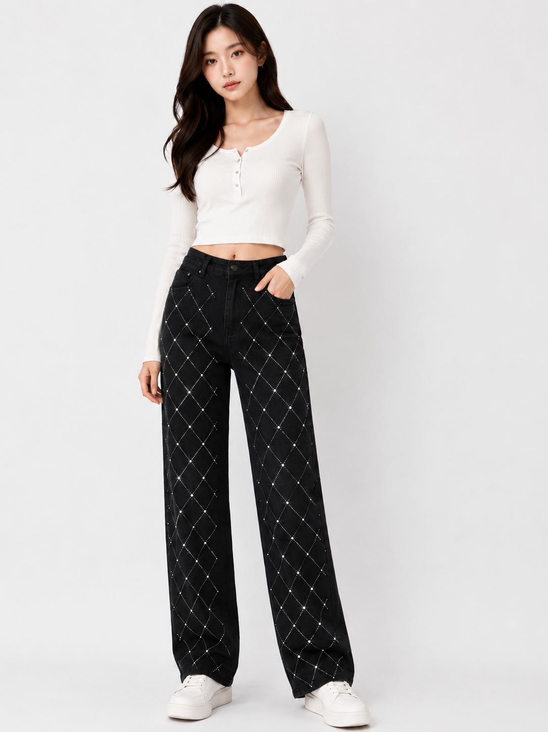 A woman modeling a white long-sleeve cropped henley top and black wide-leg pants with a white diamond lattice pattern, paired with white sneakers, against a light gray studio background.
