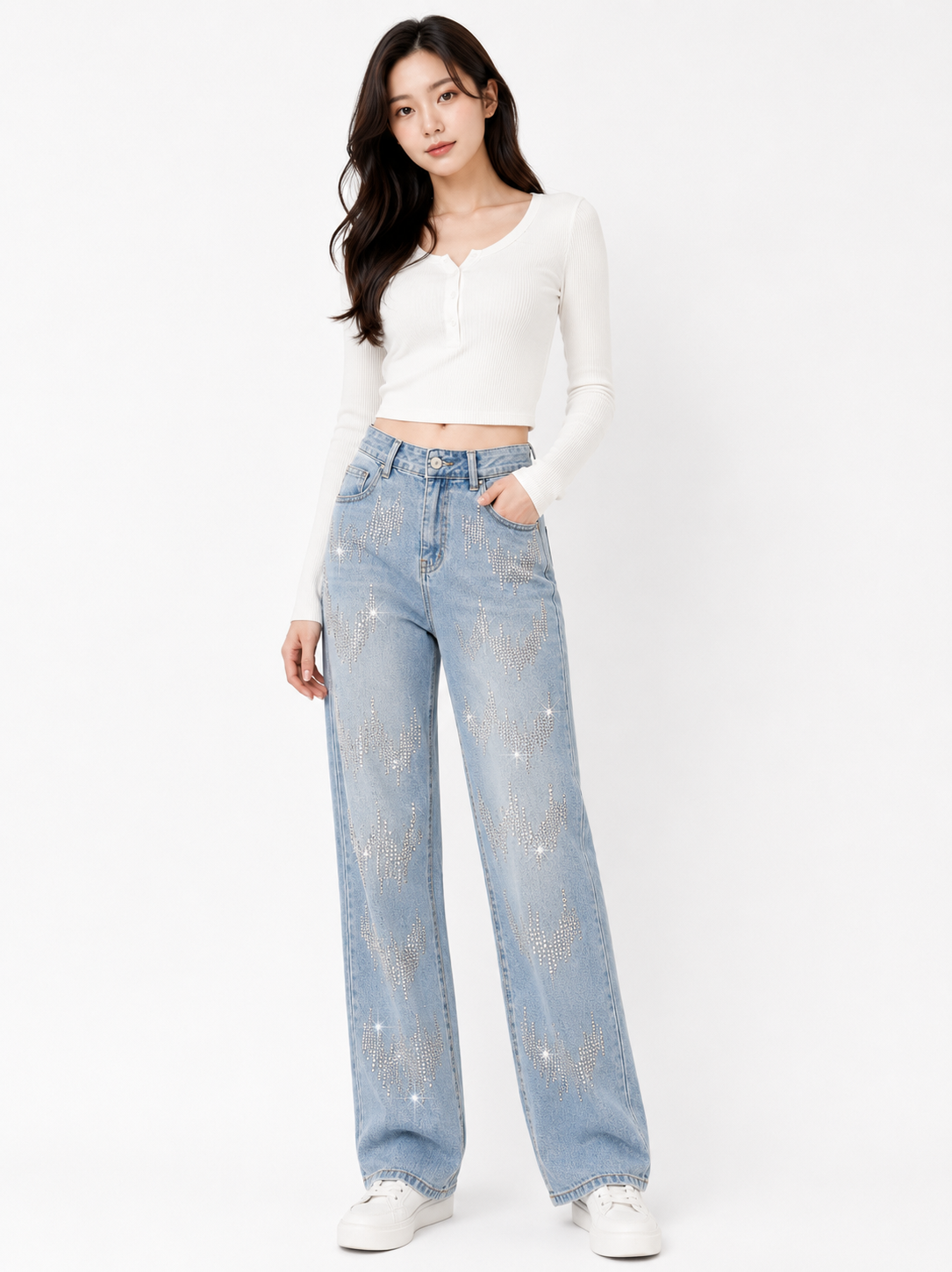 A woman modeling a pair of light-wash blue jeans with silver rhinestone embellishments, paired with a white long-sleeve crop top and white sneakers, against a plain white studio background.