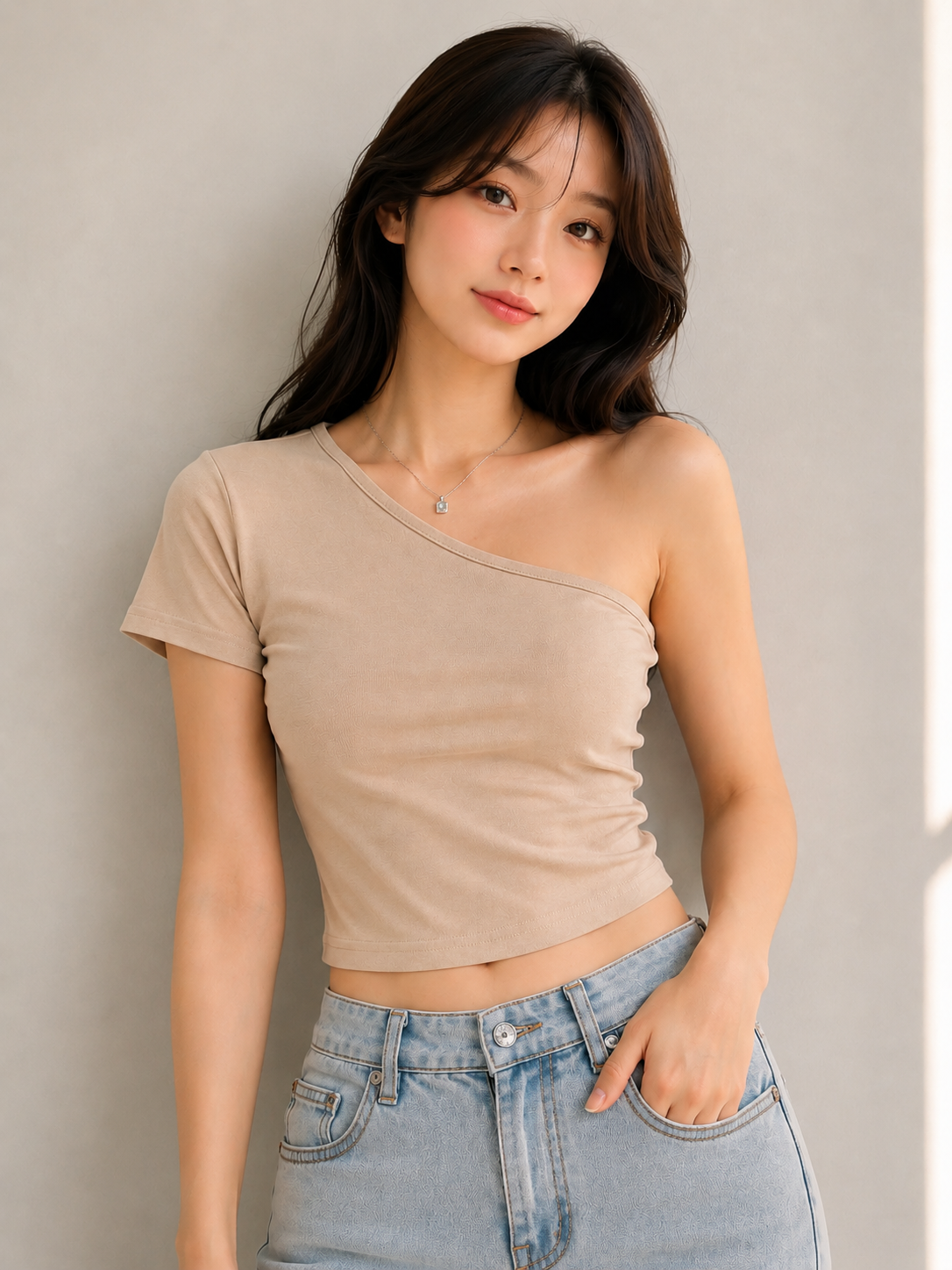A woman wearing a beige one-shoulder short-sleeve crop top and light blue jeans, standing against a neutral gray wall.
