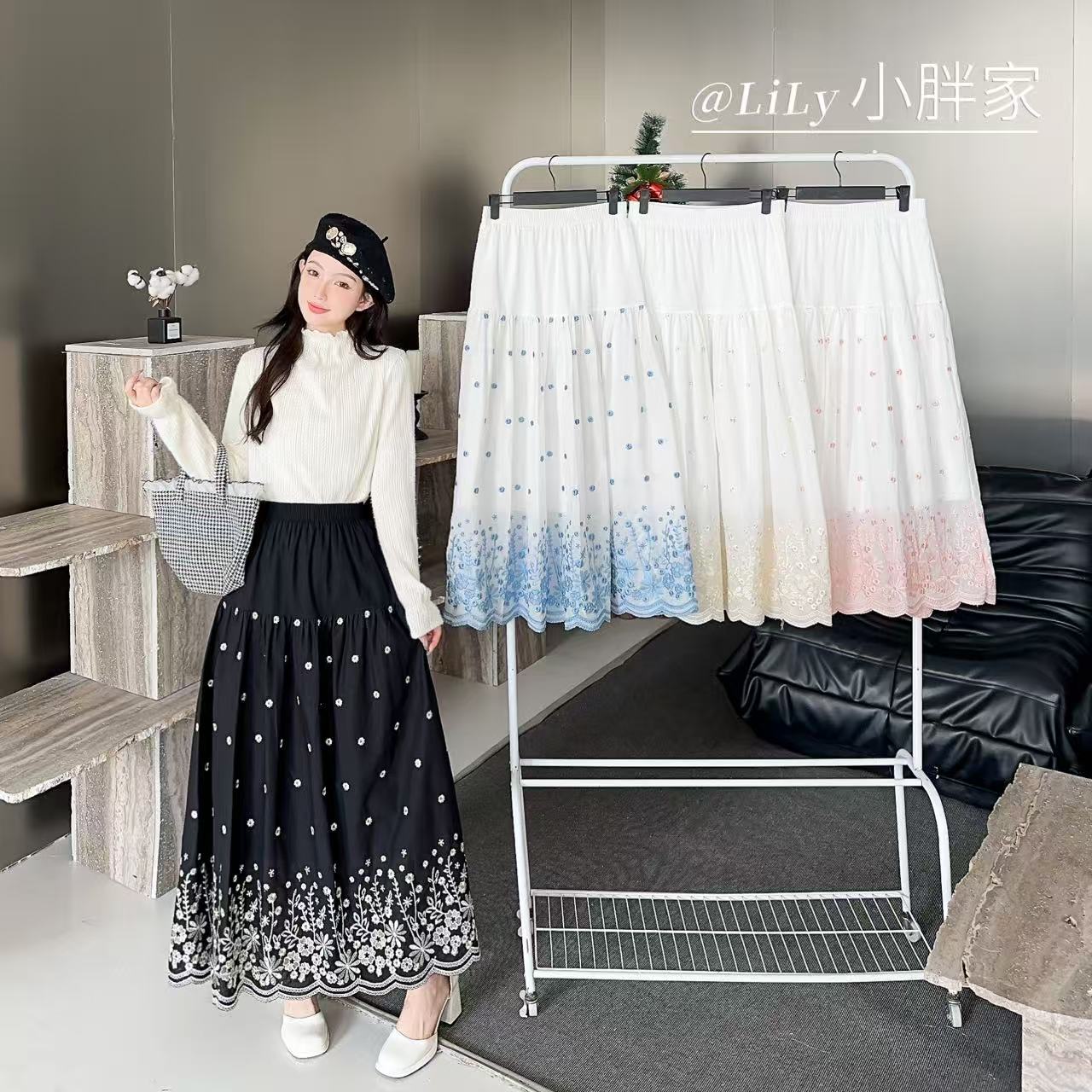 A woman models a long black skirt with white polka dots and a floral lace hem, paired with a cream mock-neck top and a black hat. To her right is a white clothing rack displaying two white tiered skirts with blue and pink gradient hem details.