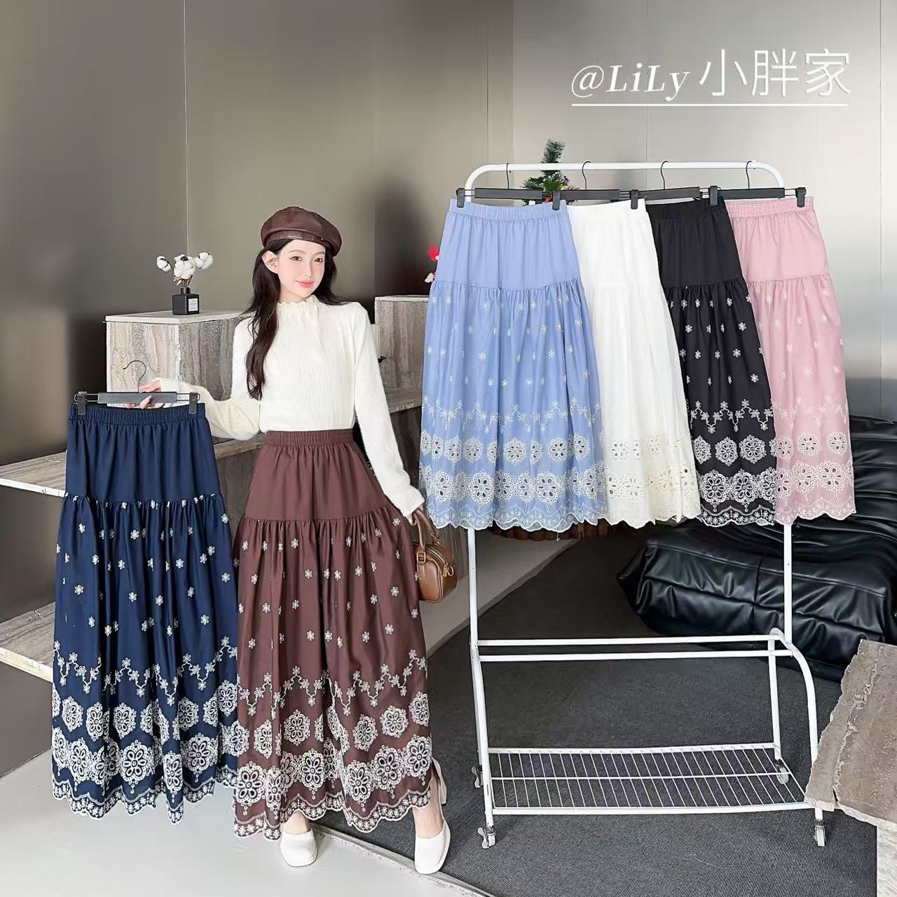 A woman models a long pleated skirt in a cream knit top and brown beret, holding up a navy blue version of the same skirt. To her right is a white clothing rack displaying multiple color options: light blue, white, black, and pink.
