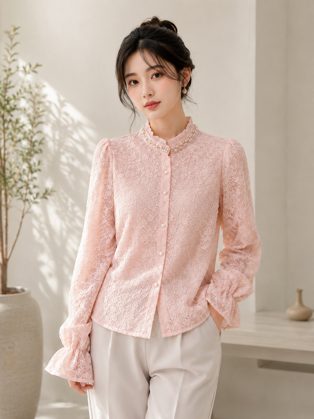 A woman wearing a light pink textured lace long-sleeve blouse with a mandarin collar and white pants, standing in a bright, modern indoor setting with soft sunlight and shadows.