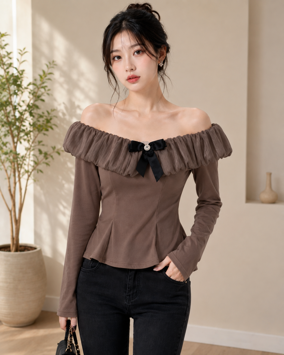A woman wearing a brown off-the-shoulder long-sleeve top with a black bow detail at the neckline, paired with black jeans, standing in a softly lit indoor setting.