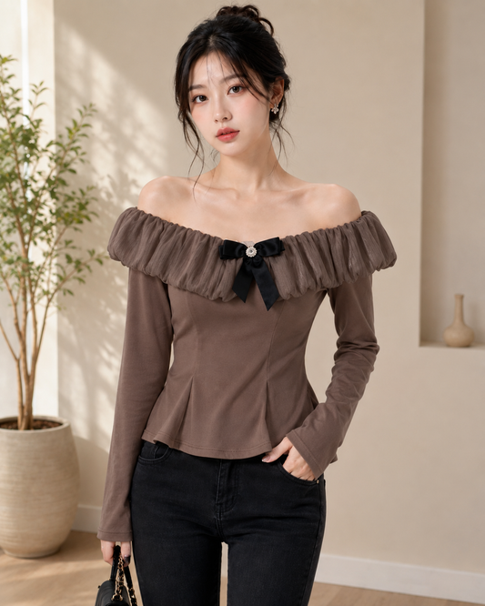 A woman wearing a brown off-the-shoulder long-sleeve top with a black bow detail at the neckline, paired with black jeans, standing in a softly lit indoor setting.