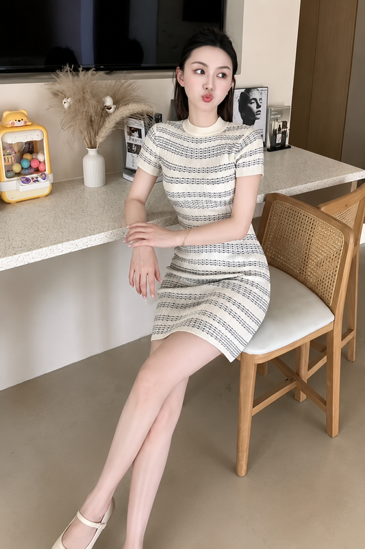 A woman wearing a short-sleeve textured knit dress with a black and white geometric pattern, sitting on a wooden chair in a modern indoor setting.