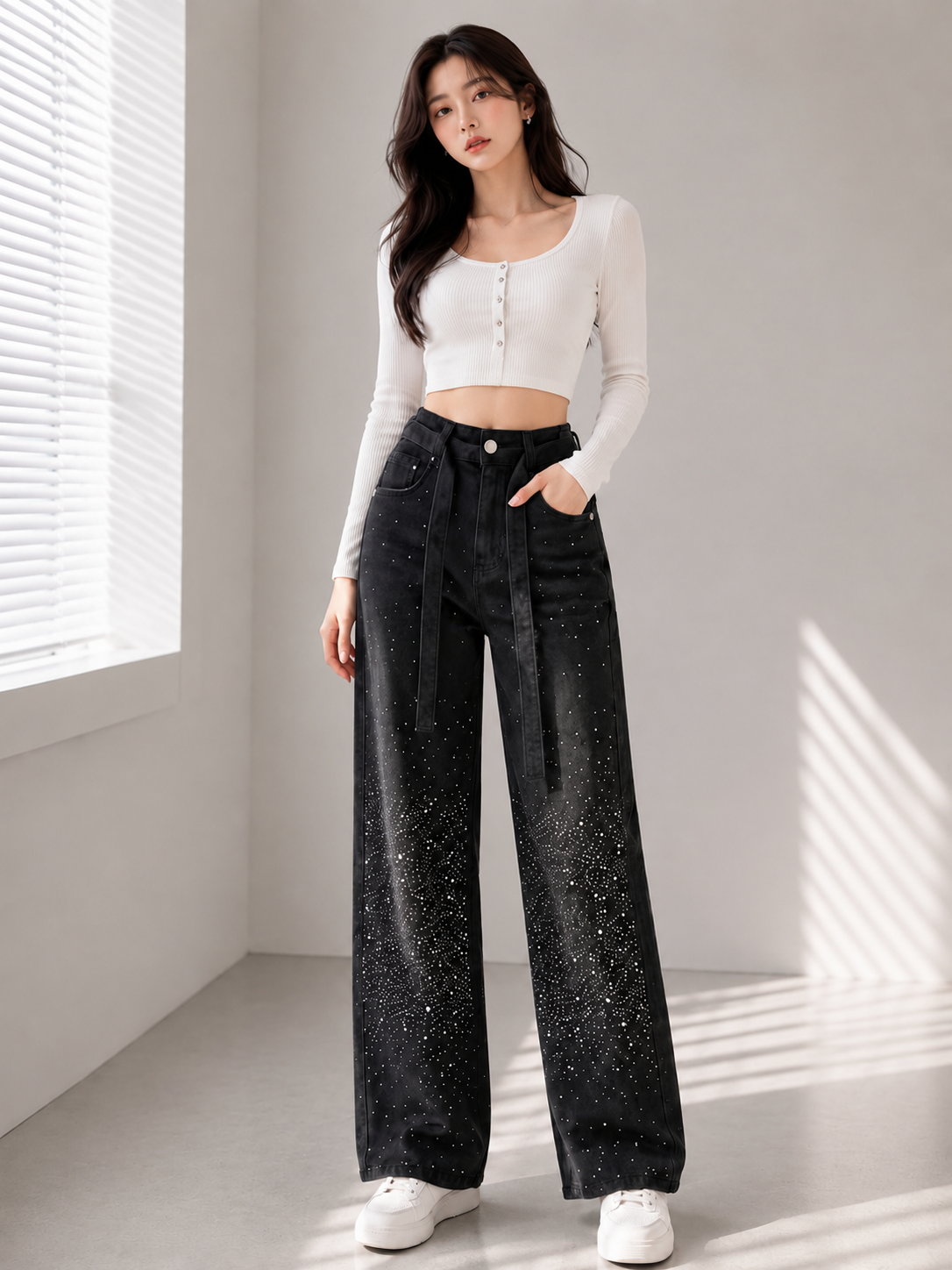 A woman stands in a bright room wearing a fitted white long-sleeve crop top, black wide-leg jeans with a subtle speckled texture, and white sneakers.