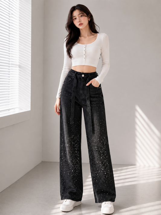 A woman stands in a bright room wearing a fitted white long-sleeve crop top, black wide-leg jeans with a subtle speckled texture, and white sneakers.