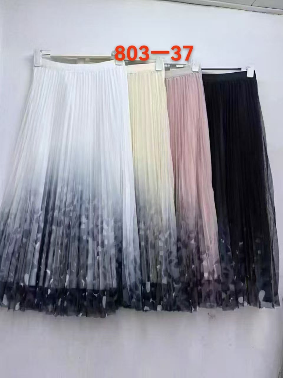 Four pleated tulle midi skirts hanging on a white wall, shown in white, cream, blush pink, and black ombre designs. An orange text overlay reads '803-37' at the top.