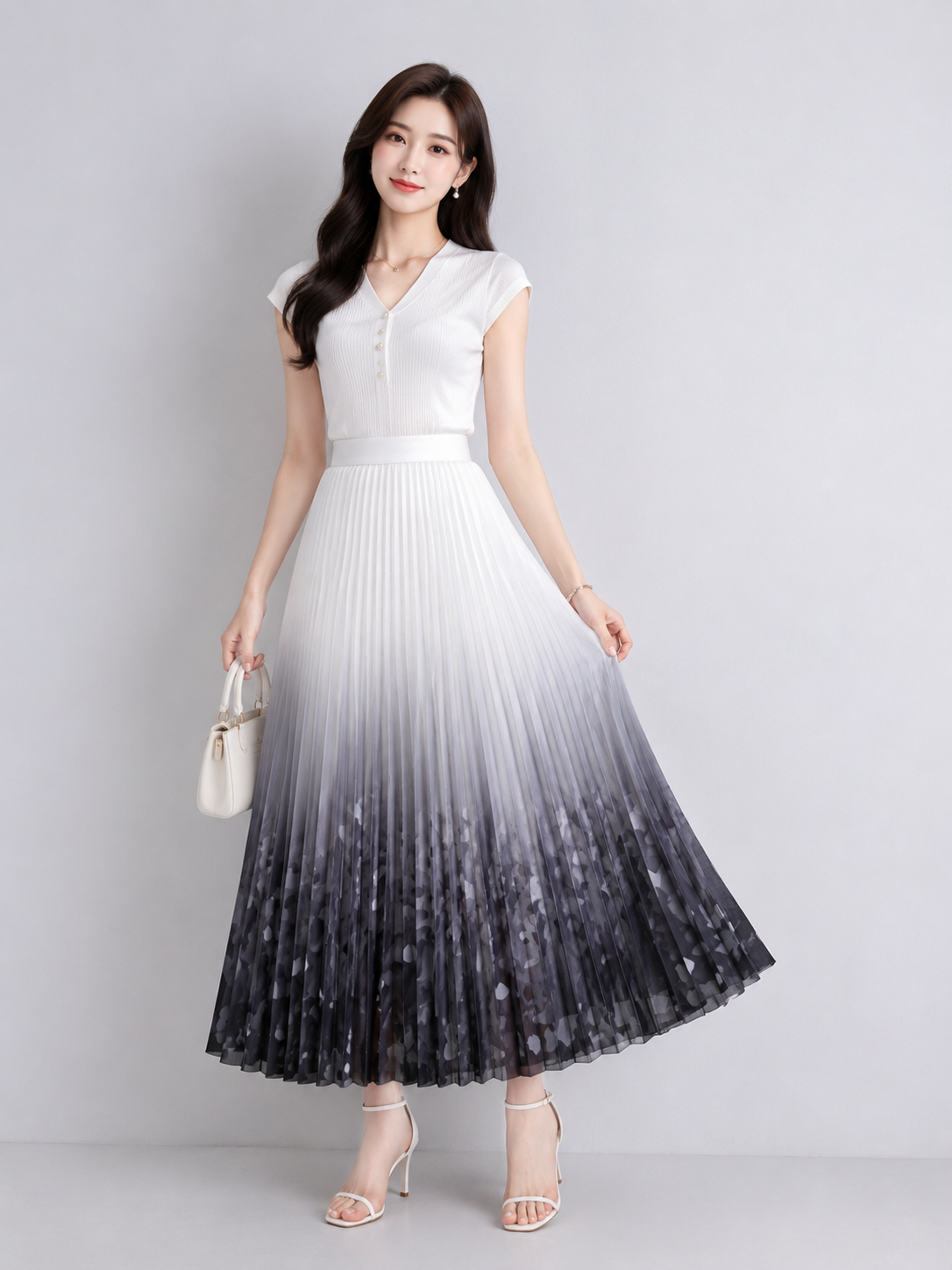 A woman models a sleeveless pleated maxi dress with a white-to-black ombre gradient design, paired with a matching waist belt and holding a small white handbag against a light gray studio background.