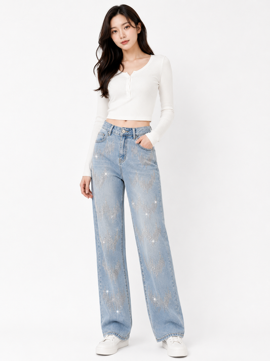 A woman modeling a pair of light-wash blue jeans with silver rhinestone embellishments, paired with a white long-sleeve crop top and white sneakers, against a plain white studio background.