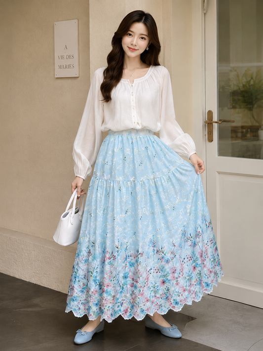 A woman stands indoors wearing a long white blouse and a light blue floral maxi skirt, holding a white handbag. The setting includes a beige wall, a door, and a small sign reading 'A VIE DES MARIES'.