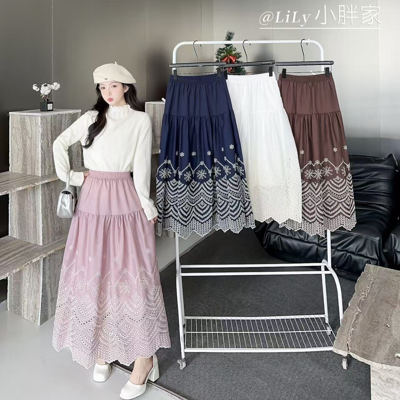 A woman models a long pleated skirt in a room setting, wearing a white beret and white top. Three other color variants of the skirt (navy blue, white, and brown) are displayed on a white clothing rack behind her.