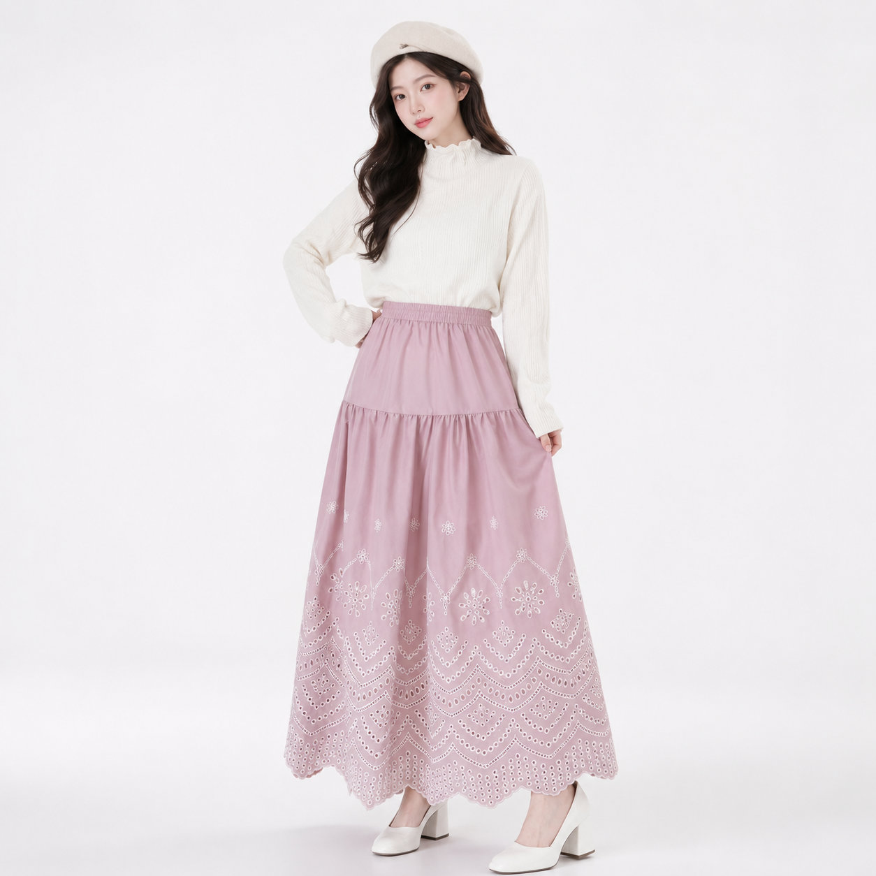 A woman modeling a long, tiered dusty pink skirt with white floral embroidery, paired with a cream mock-neck long-sleeve top, a cream beret, and white heels against a plain white studio background.