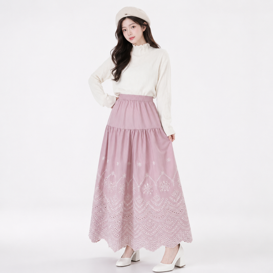 A woman modeling a long, tiered dusty pink skirt with white floral embroidery, paired with a cream mock-neck long-sleeve top, a cream beret, and white heels against a plain white studio background.