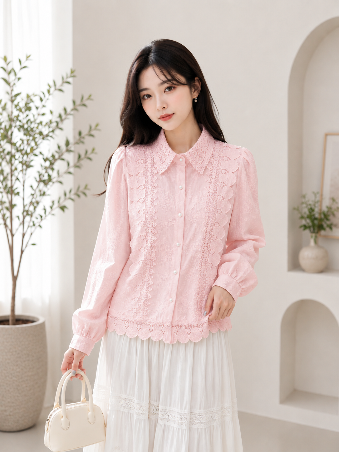 A woman wearing a light pink long-sleeve blouse with delicate lace embroidery and a scalloped hem, paired with a white skirt, holding a small cream-colored handbag in a bright, modern indoor setting.