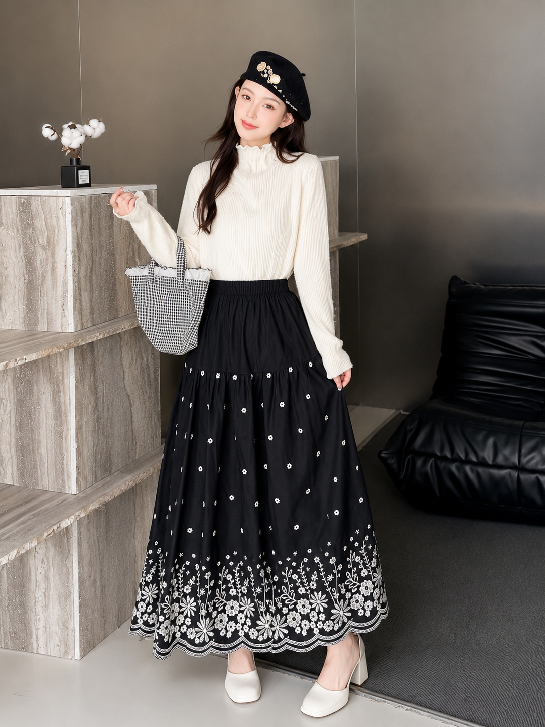 A woman stands indoors wearing a long white mock-neck top and a long black skirt with white polka dots and a white floral embroidered hem. She is also wearing a black beret and holding a gray patterned tote bag.