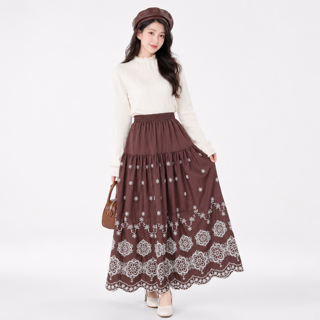 A woman modeling a long brown tiered maxi skirt with white floral embroidery and lace trim, styled with a cream mock-neck sweater, a brown beret, and a brown handbag against a plain white studio background.