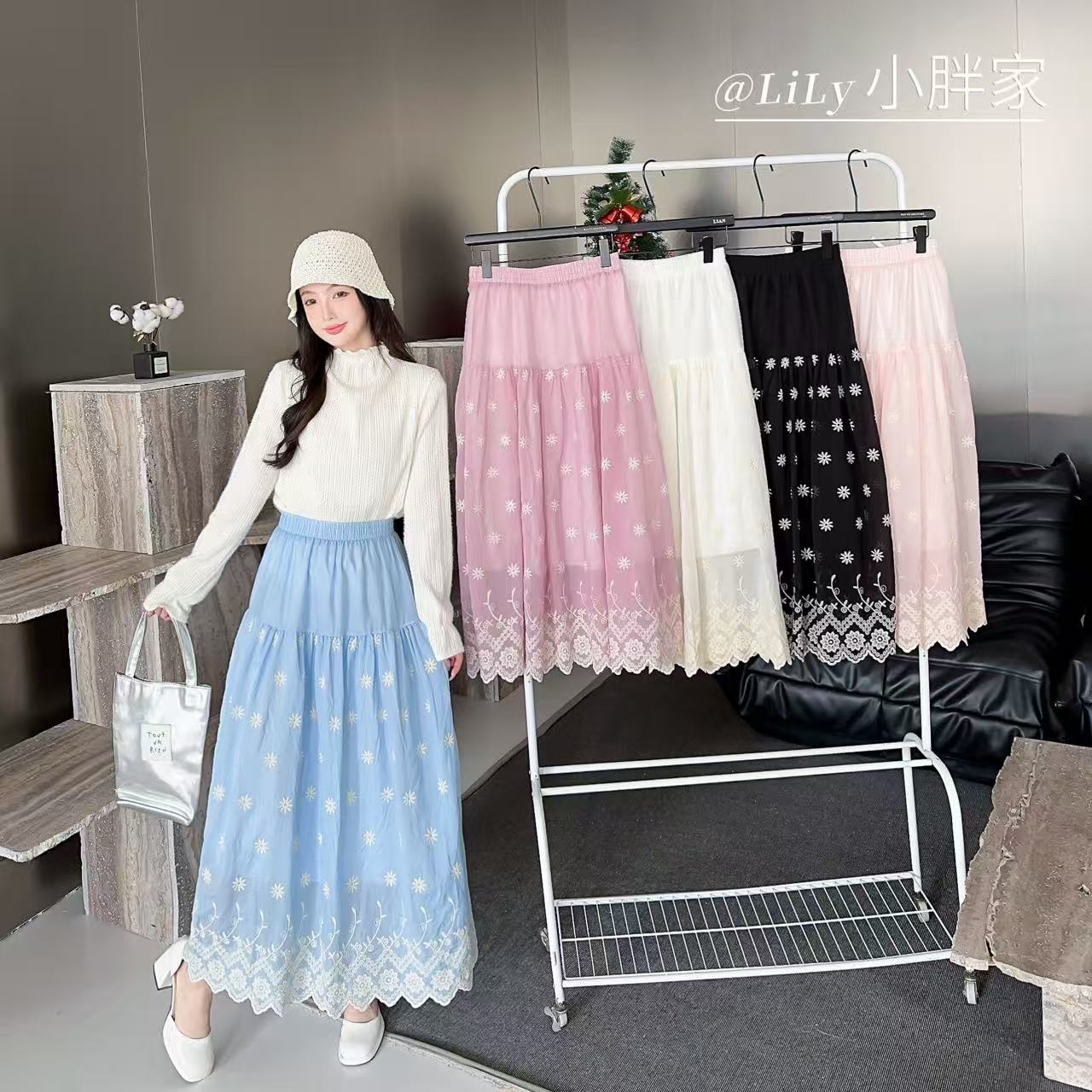 A woman models a long light blue skirt with white polka dots and a white lace hem, while standing beside a clothing rack displaying four different skirt colors: pink, white, black, and pale pink. She is wearing a white long-sleeve mock-neck top, a white bucket hat, and white shoes, holding a silver handbag.
