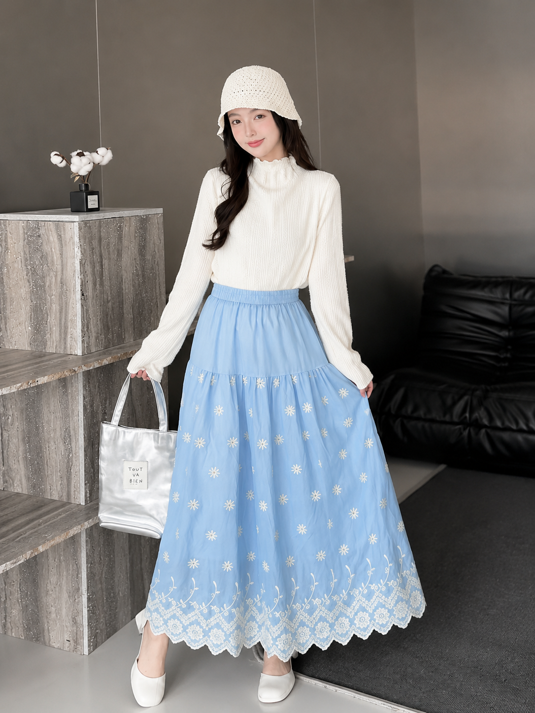 A woman models a light blue tiered maxi skirt with white floral embroidery and a scalloped lace hem, paired with a white mock-neck long-sleeve top, a white knit bucket hat, and white shoes. She is holding a white tote bag in a modern indoor setting.