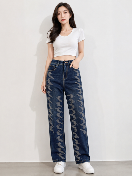 A woman modeling a pair of high-waisted, wide-leg dark blue jeans with a distinctive gray leaf-like pattern running down the front, paired with a white crop top and white sneakers against a plain white wall.